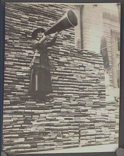 Photograph shows a woman standing on a pile of books speaking into a megaphone for an American Library Association War Service promotion to collect books for soldiers fighting in Europe.