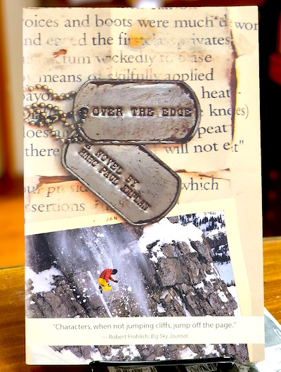 Book cover: Over The Edge: A Novel by Marc Paul Kaplan, with weathered dogs tags, old and torn printed page and photograph of a skier in mid-jump.